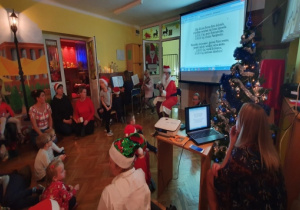 Jedna mama gra na pianinie kolędy, rodzice z dziećmi i Mikołajem wspólnie śpiewają kolędy. Tekst kolęd wyświetlany jest na ekranie.